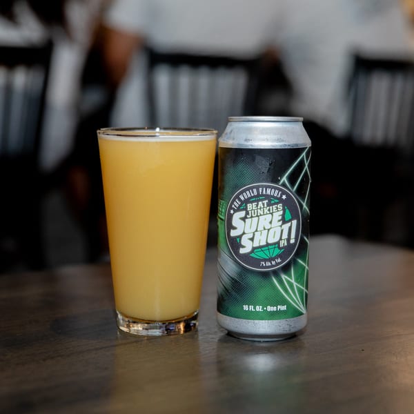 A glass of hazy IPA next to a "Beat Junkies Sure Shot!" IPA can on a wooden table.
