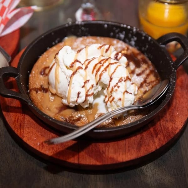 Warm skillet cookie topped with melting vanilla ice cream, caramel, and chocolate drizzle, served on a wooden board.